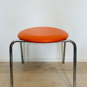 Puede incluir: Un taburete redondo con un cojín de asiento acolchado de color naranja vibrante, sostenido por cuatro patas delgadas de metal cromado pulido que se curvan en la parte superior para unirse a la base del asiento, creando un mueble funcional.