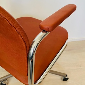 May include: A brown leather office chair with chrome arms and a swivel base. The chair has five wheels and is on a wooden floor.