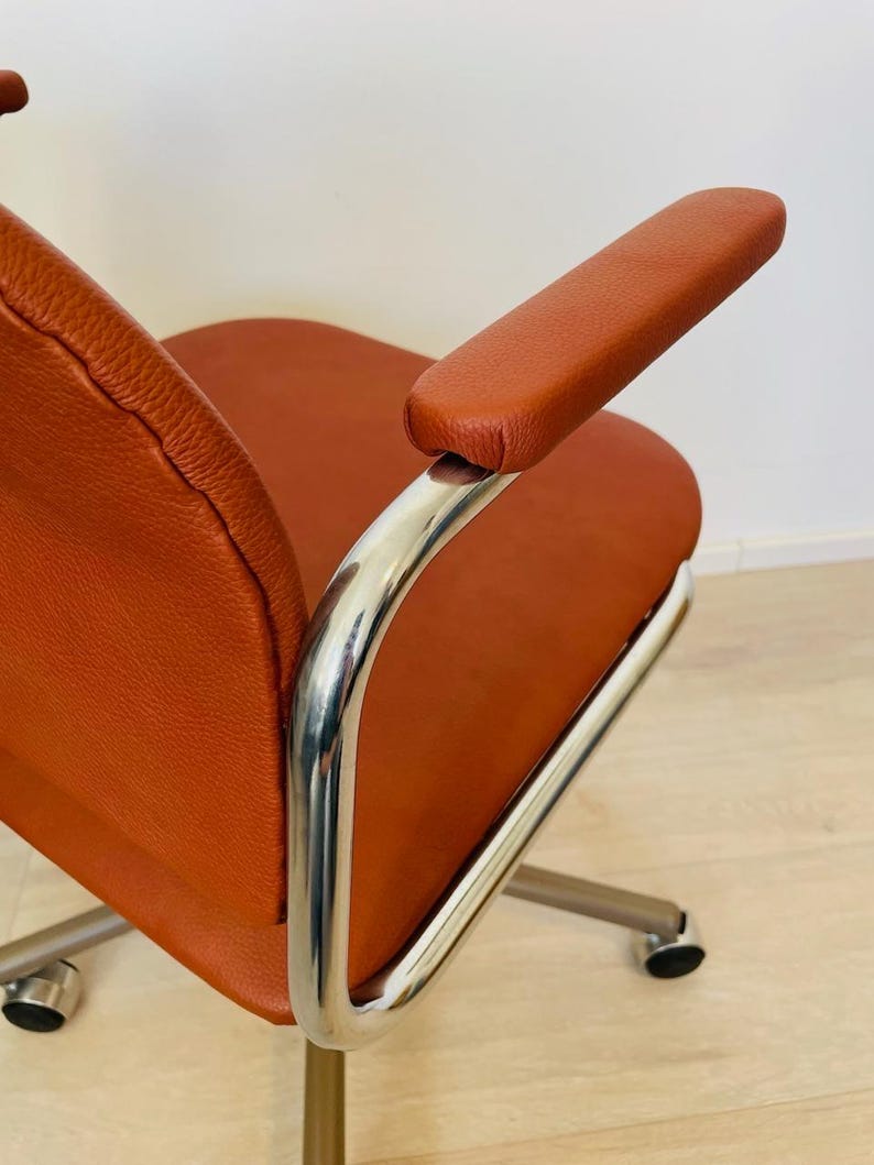 May include: A brown leather office chair with chrome arms and a swivel base. The chair has five wheels and is on a wooden floor.