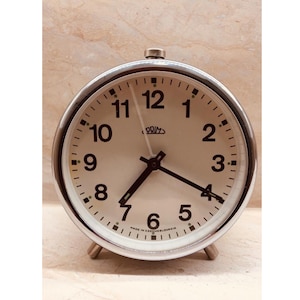 May include: A silver alarm clock with a white face and black numbers. The clock has a chrome finish and is made in Czechoslovakia. The clock is set to 7:00.