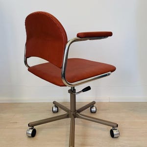 May include: A red office chair with a chrome frame and five wheels. The chair has a padded seat and back, and a single armrest on the right side.