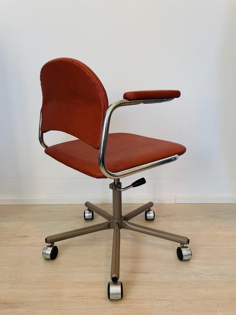 May include: A red office chair with a chrome frame and five wheels. The chair has a padded seat and back, and a single armrest on the right side.