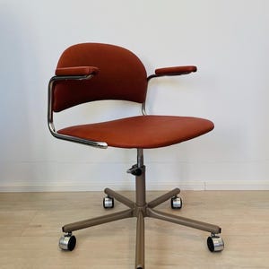 May include: A red office chair with chrome arms and a swivel base with five wheels. The chair has a padded seat and back.
