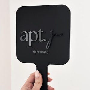 Puede incluir: Un espejo de mano negro con forma rectangular y un mango. El espejo presenta el texto "apt. j" en una fuente elegante, con el identificador de Instagram "@insideaptj" debajo.