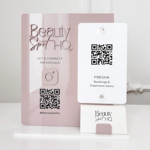 Op de afbeelding: Een roze acrylbord met de tekst "Beauty Spot HQ" en een QR-code. Een kleinere witte kaart met een QR-code en de tekst "FRESHA Bookings & Treatment menu" is bevestigd. Een visitekaarthouder met visitekaartjes is ook aanwezig.