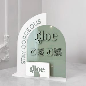 Puede incluir: Un tarjetero blanco y verde salvia con la palabra "gloe" e iconos de redes sociales. Las palabras "Stay Gorgeous" están impresas en el lateral. El soporte es de acrílico y tiene un diseño moderno.