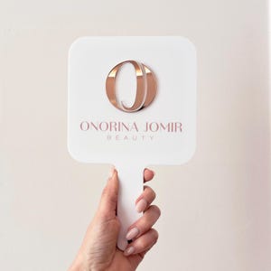 Op de afbeelding: Een witte handspiegel met een vierkante kop en handvat. De spiegel heeft een roségouden logo met de tekst "ONORINA JOMIR BEAUTY" in een zachtroze lettertype. De spiegel wordt vastgehouden door een hand met verzorgde nagels.