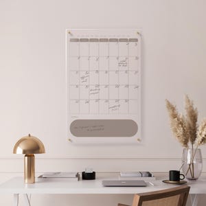 Puede incluir: Un calendario de pared de acrílico transparente con fondo blanco y diseño de cuadrícula. El calendario tiene una sección gris para notas y está montado con herrajes dorados. Se ven notas manuscritas.