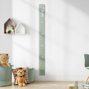 May include: A sage green height chart with white markings and the name "ELON" at the top. The chart measures up to 5 feet. The background includes children's toys and decorative items.