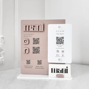 Può includere: Un espositore per un salone di bellezza, con la parola "mani" in un design 3D. Include codici QR per i social media, i pagamenti e la prenotazione di appuntamenti. L'espositore è in una combinazione di colori rosa e bianco.