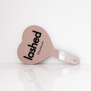 Puede incluir: Un espejo de mano en forma de corazón de color rosa pálido. La palabra "lashed" está impresa en letras negras en la superficie, con el nombre de usuario de Instagram "@lashbyem" debajo. El espejo tiene un mango con un pequeño agujero.