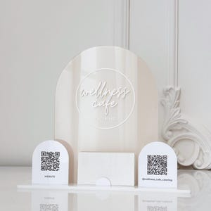 May include: A cream acrylic sign with the words "wellness cafe catering" and two QR code stands. One stand says "website" and the other says "@wellness_cafe_catering". A small white box is in the centre.