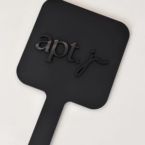 May include: A black, paddle-shaped object with a handle and a small hole at the end. The text "apt. j" is raised on the paddle's surface. The object is set against a white background.