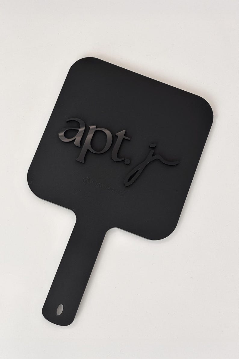 May include: A black, paddle-shaped object with a handle and a small hole at the end. The text "apt. j" is raised on the paddle's surface. The object is set against a white background.