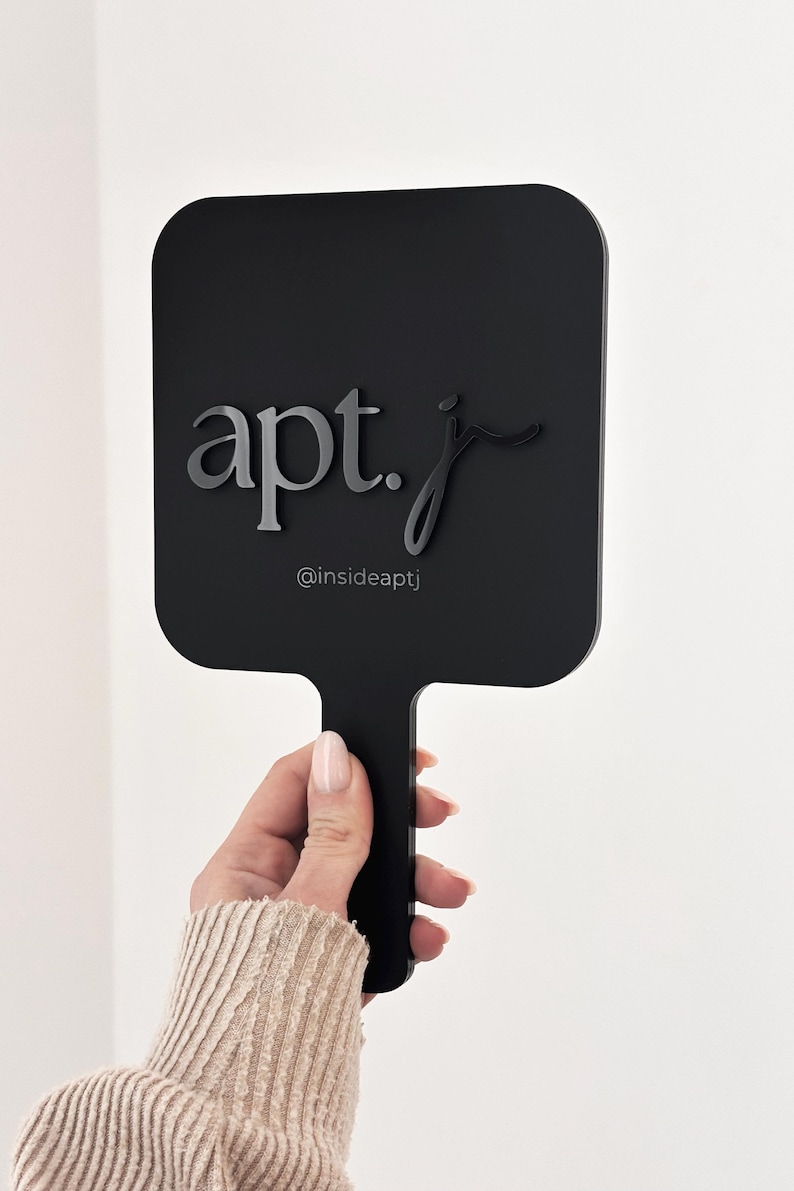 May include: A black hand mirror with a rectangular shape and a handle. The mirror features the text "apt. j" in a stylish font, with the Instagram handle "@insideaptj" below.