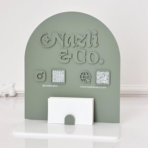 May include: A sage green business card holder with the text "Nazli & Co." and social media icons. The holder has a curved top and a slot for cards. The Instagram handle is @nazliandco and the website is www.nazliandco.com.