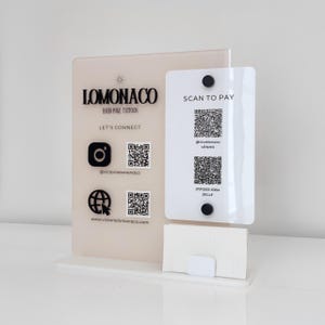 May include: A clear acrylic display stand with a light beige backing and a white front panel. The stand features the text "LOMONACO HAND POKE TATTOOS" and social media QR codes. A white card holder is at the base, and the front panel says "SCAN TO PAY".