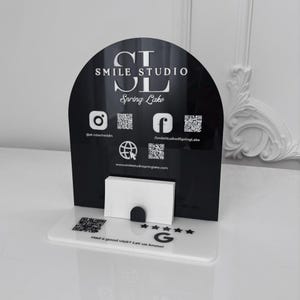 Puede incluir: Un tarjetero de acrílico en blanco y negro con el texto "SMILE STUDIO Spring Lake". El soporte tiene iconos de redes sociales y códigos QR. La base incluye el texto "Had a good visit? Let us know!"