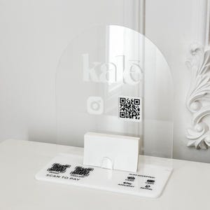 Puede incluir: Portatarjetas de visita de acrílico transparente con una parte superior arqueada. La palabra "kale" está grabada en la parte superior. Incluye un código QR y el logotipo de Instagram. La base dice "Scan to Pay" y "Also Accepted: Card, Cash, Apple Pay, PayPal."