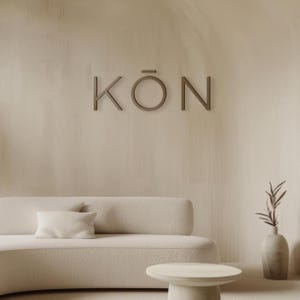 May include: A neutral-toned interior scene featuring a curved sofa, a small round table, and a decorative vase with a plant. The word "KÖN" is displayed on the wall in a minimalist font. The color palette is soft and inviting.