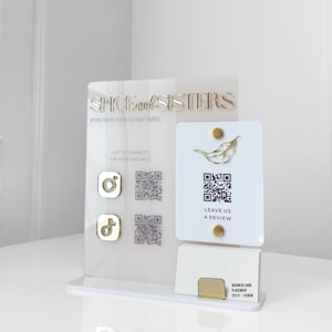 Puede incluir: Un tarjetero de acrílico transparente con detalles dorados. El soporte muestra el texto "SPICE and SISTERS" e iconos de redes sociales. Una tarjeta separada presenta un diseño de hoja y códigos QR. La base incluye un área para tarjetas de visita.