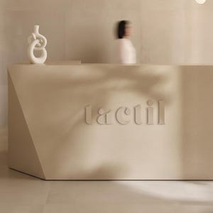 Puede incluir: Un mostrador de recepción beige con la palabra "tactil" en relieve en la parte delantera. Un jarrón decorativo blanco está sobre el mostrador. Una persona está borrosa en el fondo.