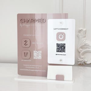 Puede incluir: Un expositor acrílico de dos partes en tonos malva y blanco. La sección más grande presenta el texto "CHARMED by Maggie" e información de contacto. La sección más pequeña tiene "LET'S CONNECT" y un código QR. Un tarjetero está en la base.