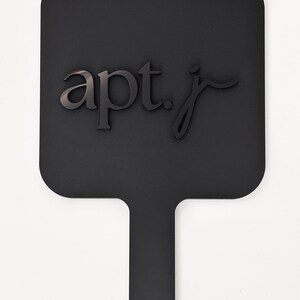 May include: A black paddle-shaped sign with rounded corners and a handle. The sign features the text "apt. j" in a dark, stylized font. A small hole is at the end of the handle. The background is plain white.