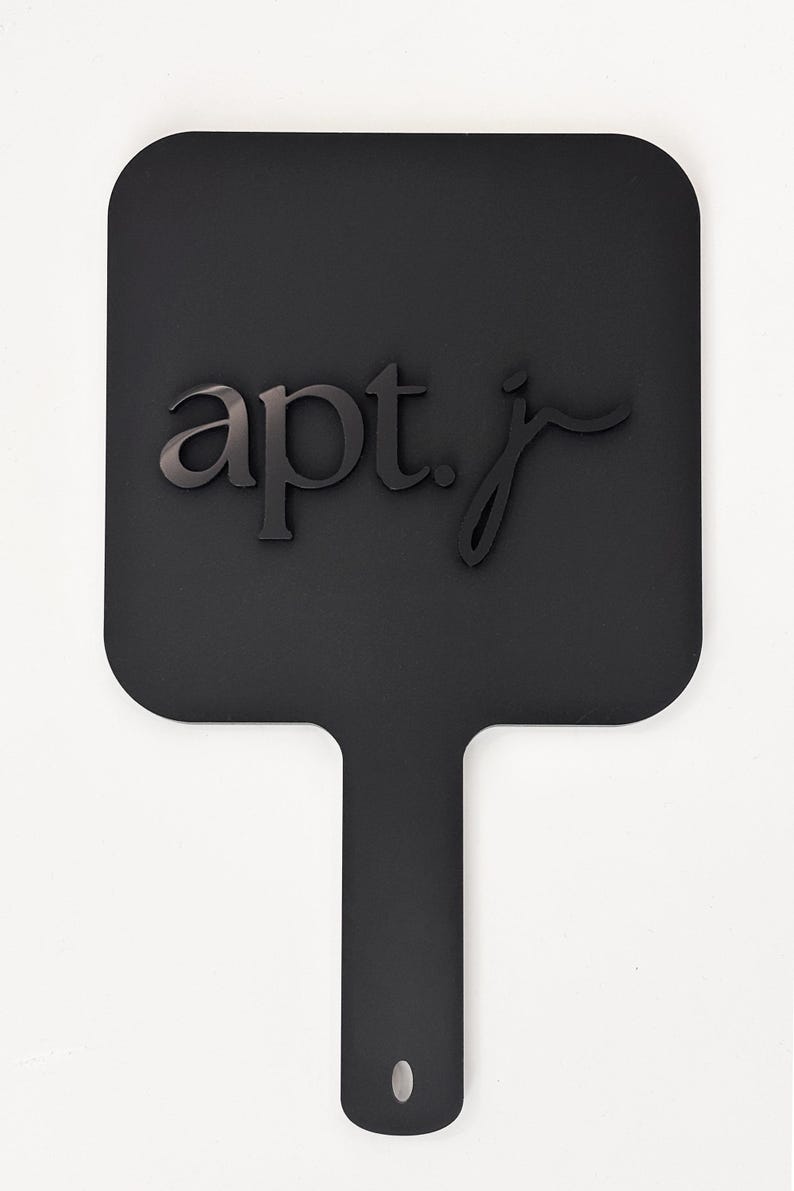May include: A black paddle-shaped sign with rounded corners and a handle. The sign features the text "apt. j" in a dark, stylized font. A small hole is at the end of the handle. The background is plain white.