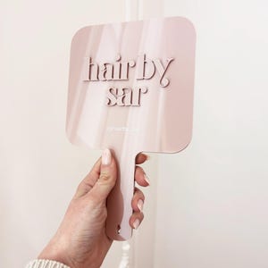 Puede incluir: Un espejo de mano rosa con el texto "hair by sar". El espejo tiene una forma rectangular con esquinas redondeadas y un mango. El texto "@hairby_sar" está en la parte inferior. El espejo lo sostiene una persona.