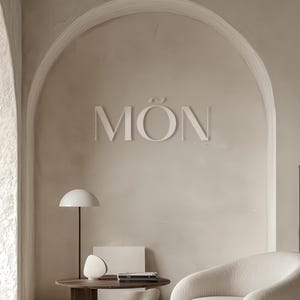 May include: A white, three-dimensional sign with the word "MÓN" on a beige wall with an arched doorway. A white lamp with a round shade sits on a wooden table next to a white book and a white armchair.