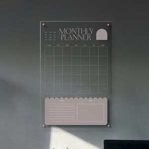 May include: A clear acrylic monthly planner with gold-colored hardware. The planner features a grid for scheduling, a section for to-do lists, and the words "MONTHLY PLANNER" in a serif font.