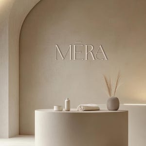 Puede incluir: Interior de tonos neutros con la palabra "MERA" en relieve en la pared. Una encimera redonda beige exhibe un cuenco pequeño, una botella, una toalla enrollada y un jarrón con plantas secas. La estética general es minimalista y de estilo spa.