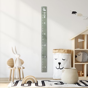 May include: A sage green height chart with white markings and the name "ELON" printed vertically. The chart measures up to 6 feet. The image shows a child's room with a toy rabbit, a wooden chair, and a bear-themed storage bag.
