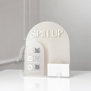Può includere: Un porta biglietti da visita in acrilico color crema con la scritta "SPIN UP POLE DANCE & AERIAL ARTS". Presenta un pannello più piccolo con codici QR dei social media e le parole "LET'S CONNECT" e "SCAN TO PAY".