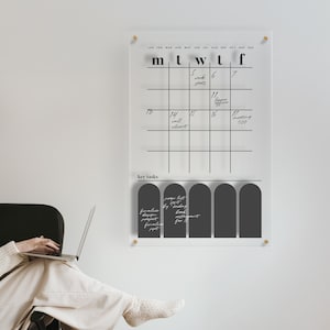 May include: A clear acrylic wall calendar with a grid layout and the days of the week labeled across the top. The calendar has handwritten notes and a section for key tasks. Gold hardware secures the calendar to the wall.