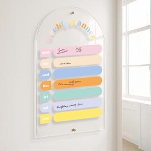 May include: Clear acrylic weekly planner with a curved top. The words "weekly planner" are in pastel colours. The days of the week are in pastel colours. Each day has a pastel coloured rectangular section for notes. Includes gold-coloured hardware.