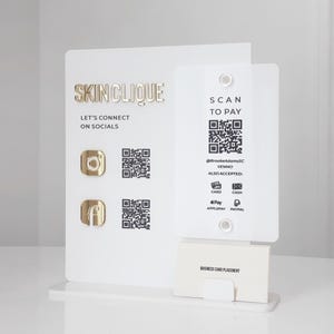 Puede incluir: Un expositor de acrílico blanco con letras doradas "SKINCLIQUE". El expositor tiene una sección para tarjetas de visita y códigos QR para pagos y enlaces de redes sociales. También es visible el texto "SCAN TO PAY".