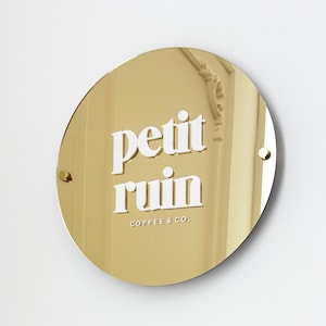 Puede incluir: Un letrero redondo dorado con las palabras "petit ruin" en blanco y negrita. Debajo, el texto "COFFEE & CO." también está en blanco. El letrero está fijado a una pared blanca con dos pequeños tornillos dorados.