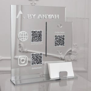 Puede incluir: Expositor de acrílico transparente con el texto "BY ANYAH", iconos de redes sociales y códigos QR. El expositor incluye un tarjetero y un letrero más pequeño con el texto "Order here:" y un código QR.