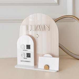 Puede incluir: Un tarjetero de acrílico color crema con la palabra "LUMIN" en letras doradas. El soporte tiene una parte superior curvada y un pequeño acento de estrella. Una sección blanca más pequeña muestra iconos de redes sociales y el texto "LET'S CONNECT".