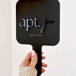 May include: A black hand mirror with a square shape and a handle. The mirror features the text "apt. j" in a stylized font, with the Instagram handle "@insideaptj" below. The mirror is held against a white background.