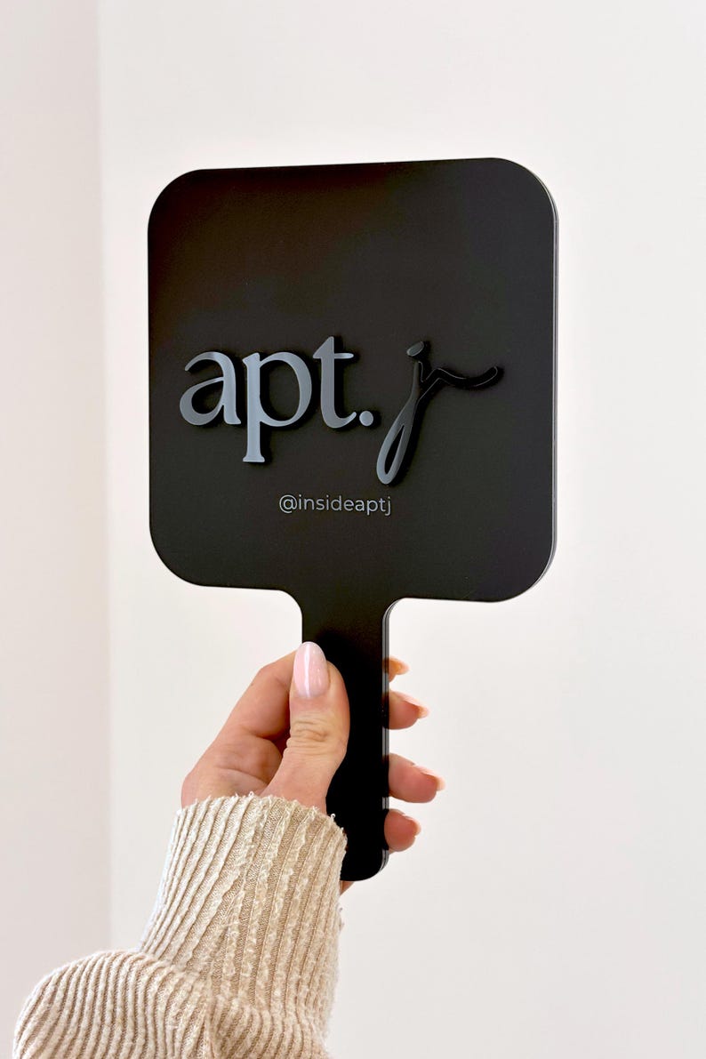 May include: A black hand mirror with a square shape and a handle. The mirror features the text "apt. j" in a stylized font, with the Instagram handle "@insideaptj" below. The mirror is held against a white background.