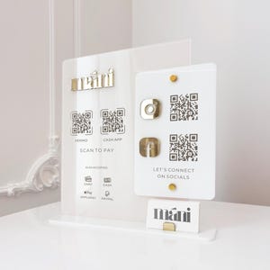Puede incluir: Un letrero de negocios de acrílico transparente con detalles dorados. El letrero muestra códigos QR para pago y redes sociales, junto con el texto "VENMO", "CASH APP", "SCAN TO PAY" y "LET'S CONNECT ON SOCIALS". Una tarjeta de visita con el logo "mani" está en un soporte dorado.