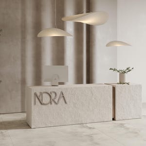 Könnte beinhalten: Ein moderner Empfangstresen mit dem Namen "NORA" in silbernen Buchstaben. Der Tresen besteht aus einem strukturierten, hellbeigen Material. Drei einzigartige, geschwungene Pendelleuchten hängen darüber. Ein Computermonitor und eine kleine Vase mit Grün sind ebenfalls vorhanden.