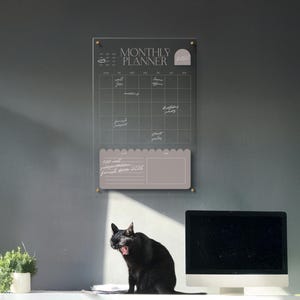 Puede incluir: Un planificador mensual de acrílico transparente con el texto "MONTHLY PLANNER 2020". El planificador tiene una cuadrícula para programar y una sección separada para notas. Un gato negro bosteza frente a un monitor de computadora.