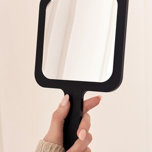 May include: A black, square-shaped hand mirror with rounded corners. The mirror is held by a black handle. The frame is black, and the reflective surface is clear. The mirror is held in front of a neutral background.
