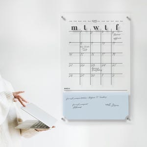 Puede incluir: Un calendario de pared de acrílico transparente con una cuadrícula negra y notas manuscritas. El calendario tiene una sección de notas azul claro en la parte inferior. Una persona sostiene un ordenador portátil en primer plano.