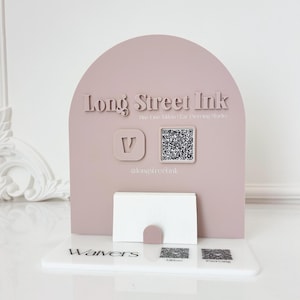 Puede incluir: Un tarjetero de color malva con el texto "Long Street Ink" y un código QR. El soporte tiene una parte superior curvada y una ranura para tarjetas. La base tiene la palabra "Waivers" y dos códigos QR adicionales.