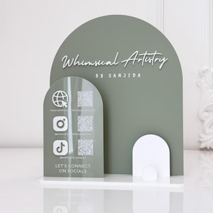 Può includere: Un espositore in acrilico verde salvia e bianco con il testo "Whimsical Artistry" e icone di social media. Lo stand include una base e una parte superiore curva, con le parole "Let's Connect on Socials".